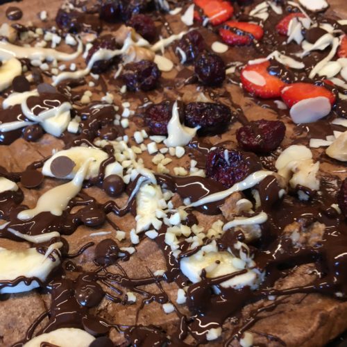 Schokoladiges Brownie Blech von oben fotografiert mit vielen Toppings aus Erdbeeren, Nüssen, Schokostücken und Nussmus.