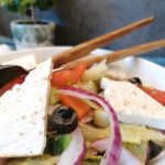 Griechischer Salat angerichtet in einem tiefen weißen Teller. In der Schale sind von rechts ein großes Salatbesteck. Im Hintergrund steht eine Pflanze in einem grauen Topf.